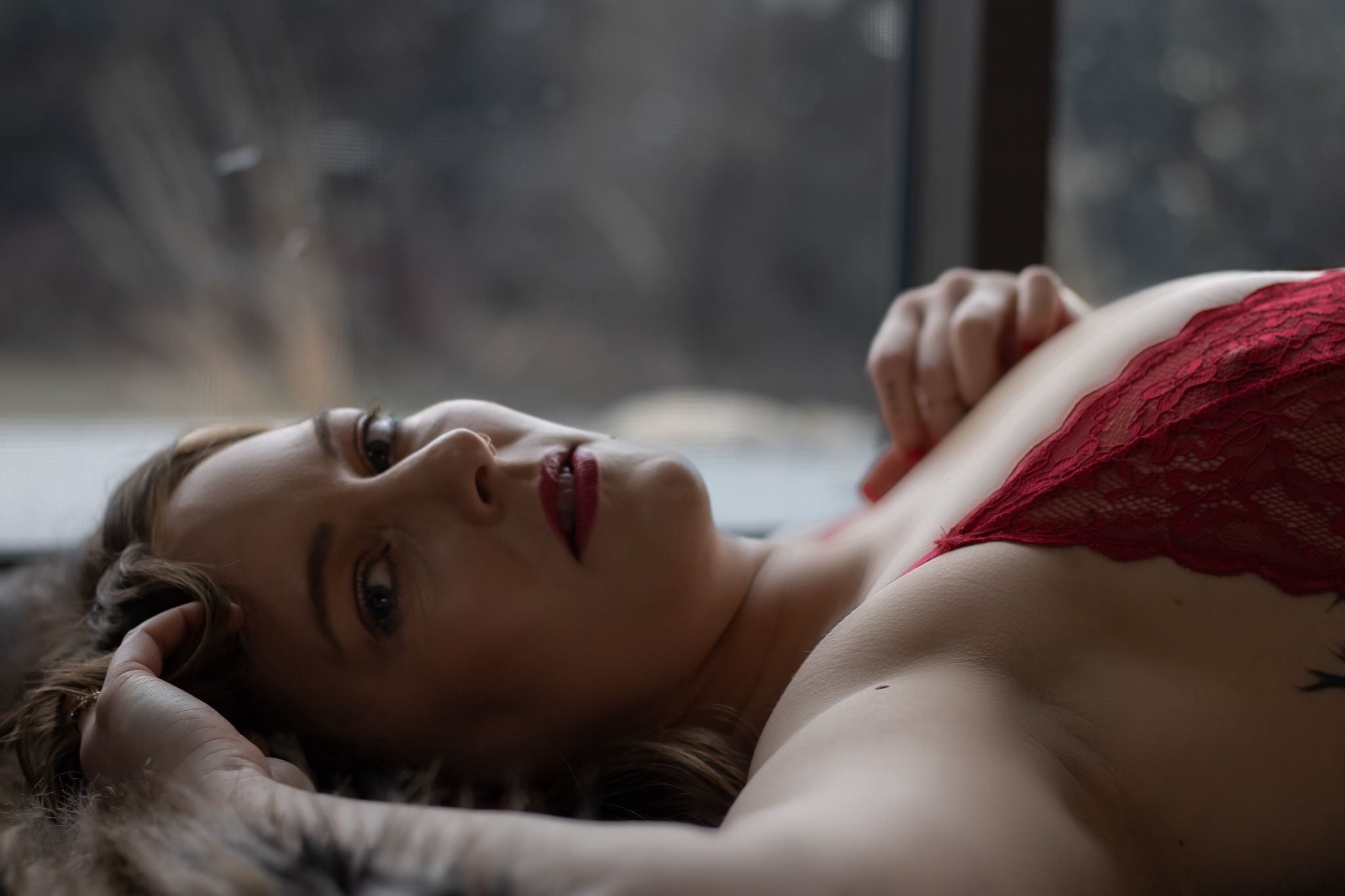 Sensual portrait of woman in red lingerie lying on fur with dreamy window light.
