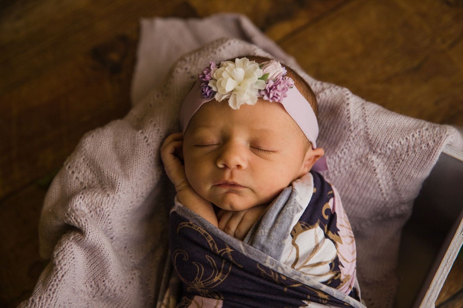 Newborn wearing floral headband sleeping peacefully on purple blanket with hardwood floor background.
