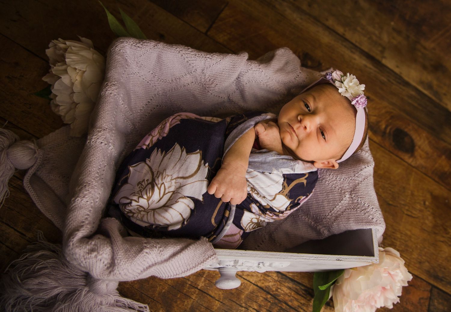 Sleeping newborn in lace headband and floral wrap on purple blanket with wooden backdrop.
