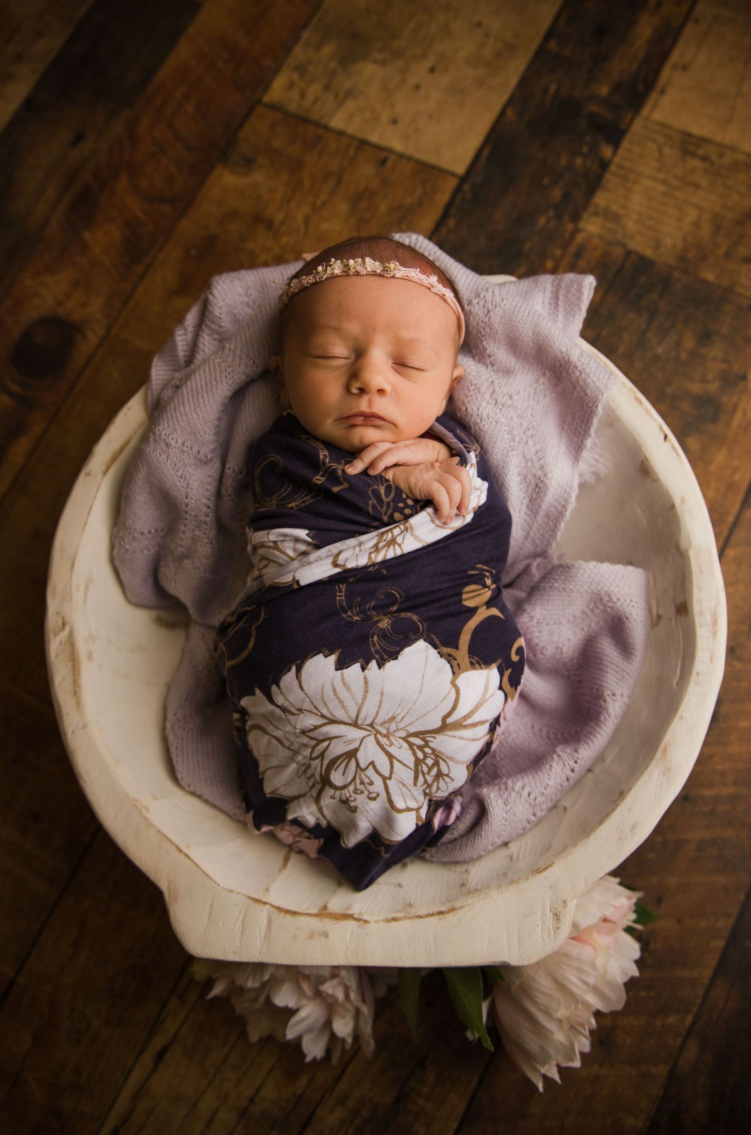 Peaceful sleeping newborn in floral wrap and headband nestled in white bowl on wooden floor.