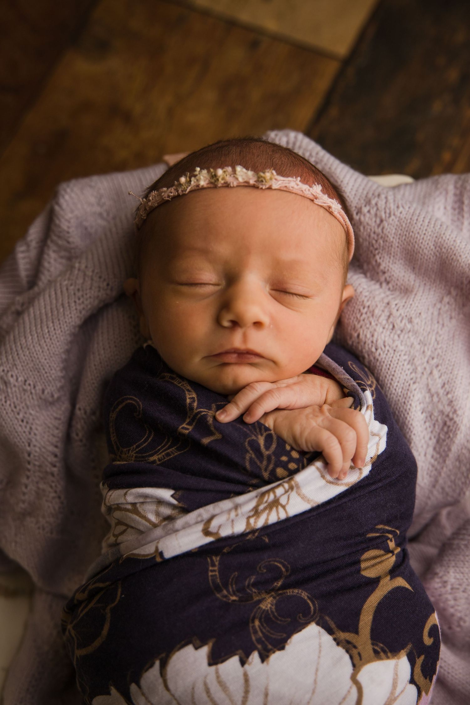 Peaceful sleeping newborn with headband and navy floral swaddle on purple blanket.