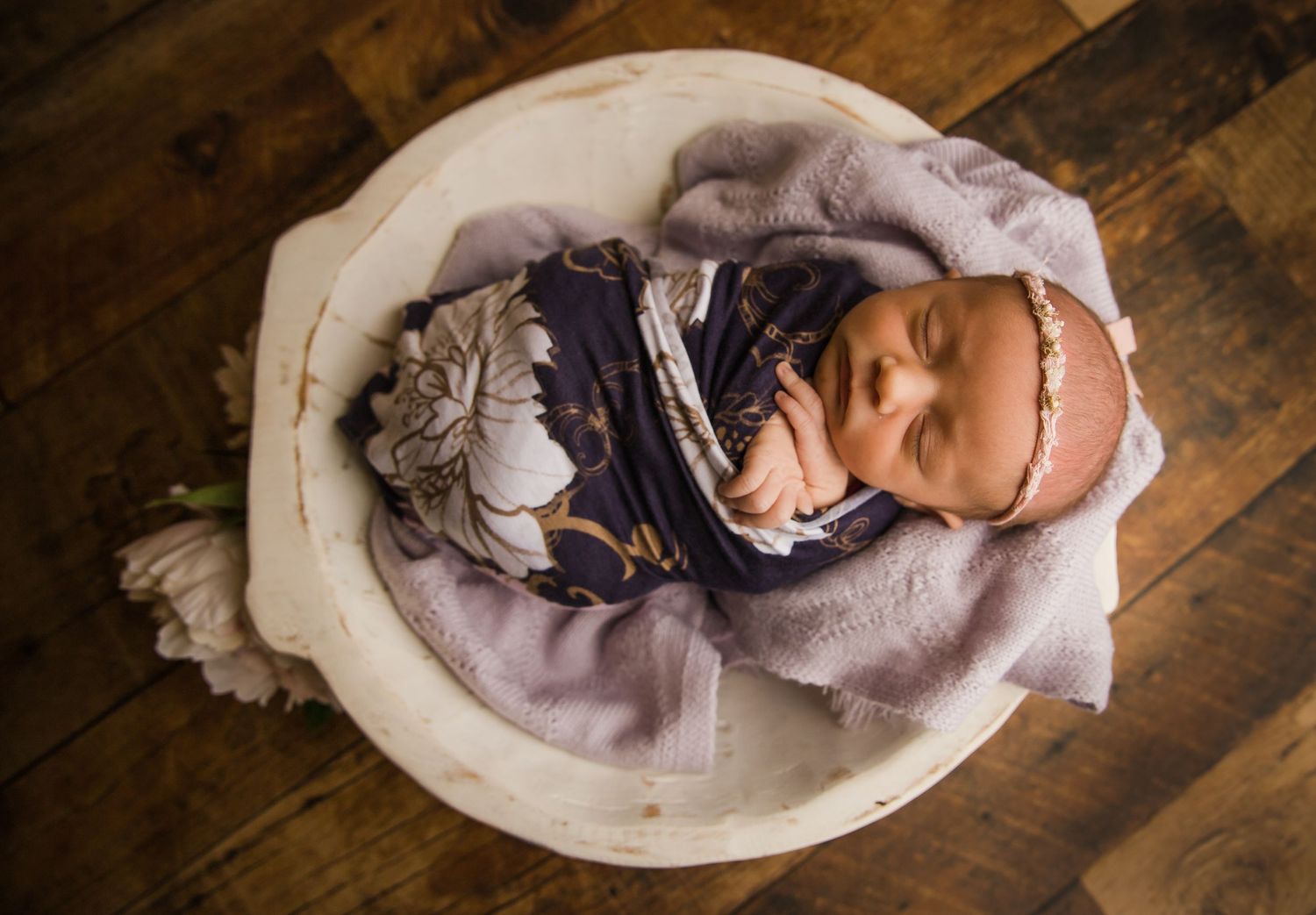 Overhead view of sleeping newborn in white bowl with purple blanket and floral wrap.