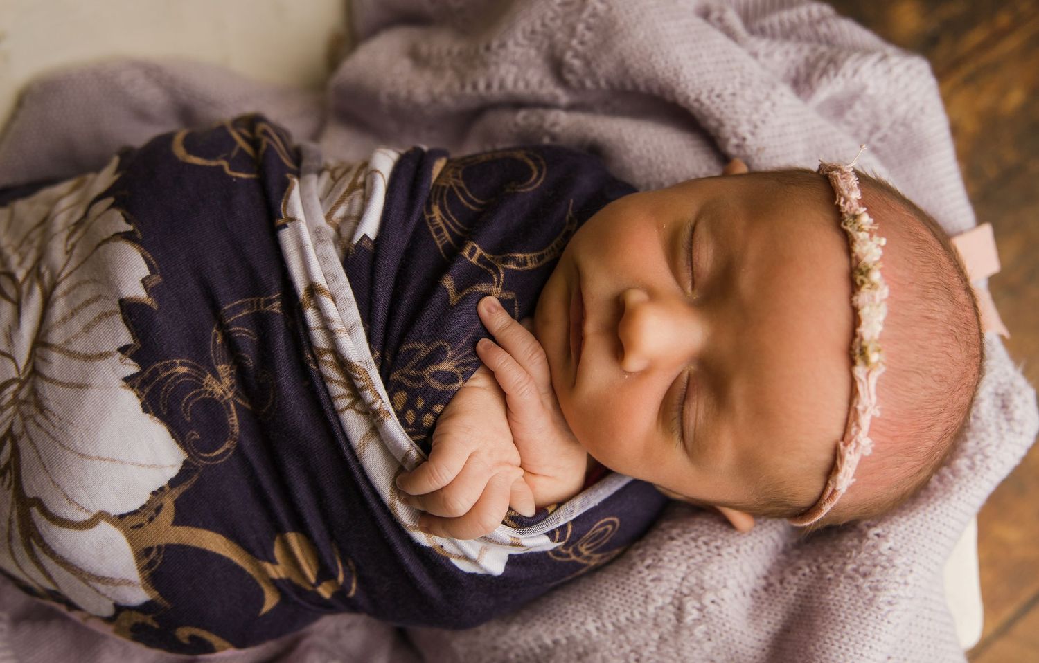 A sleeping baby wearing a navy floral outfit and delicate lace headband rests on soft fabric.