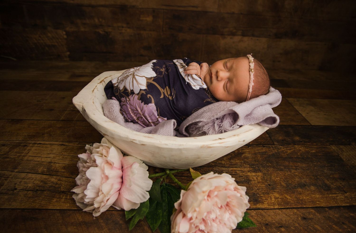 A newborn rests in a white basket with pink peonies on wooden surface.