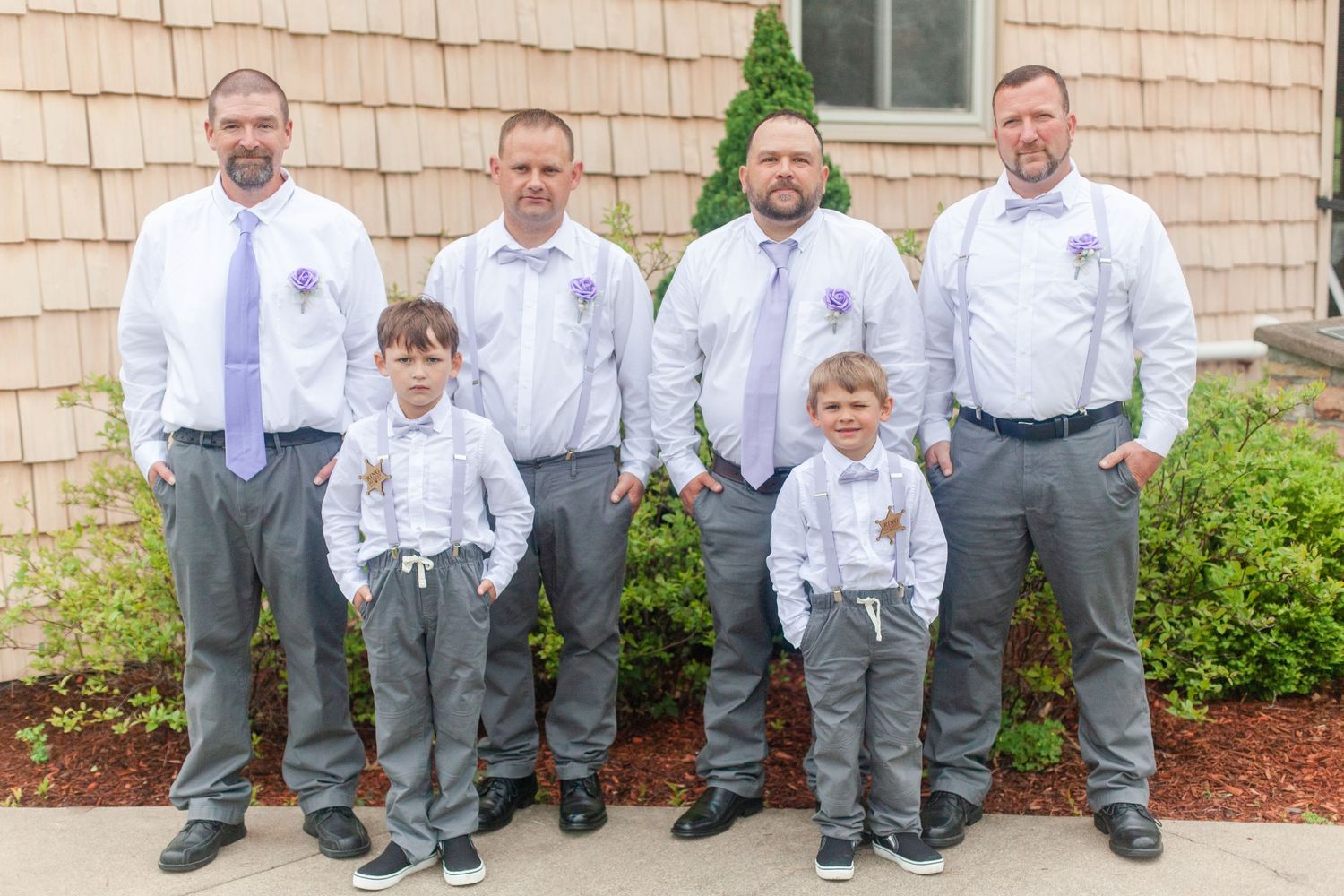 Groomsmen in white shirts and gray pants lined up against beige building exterior.
