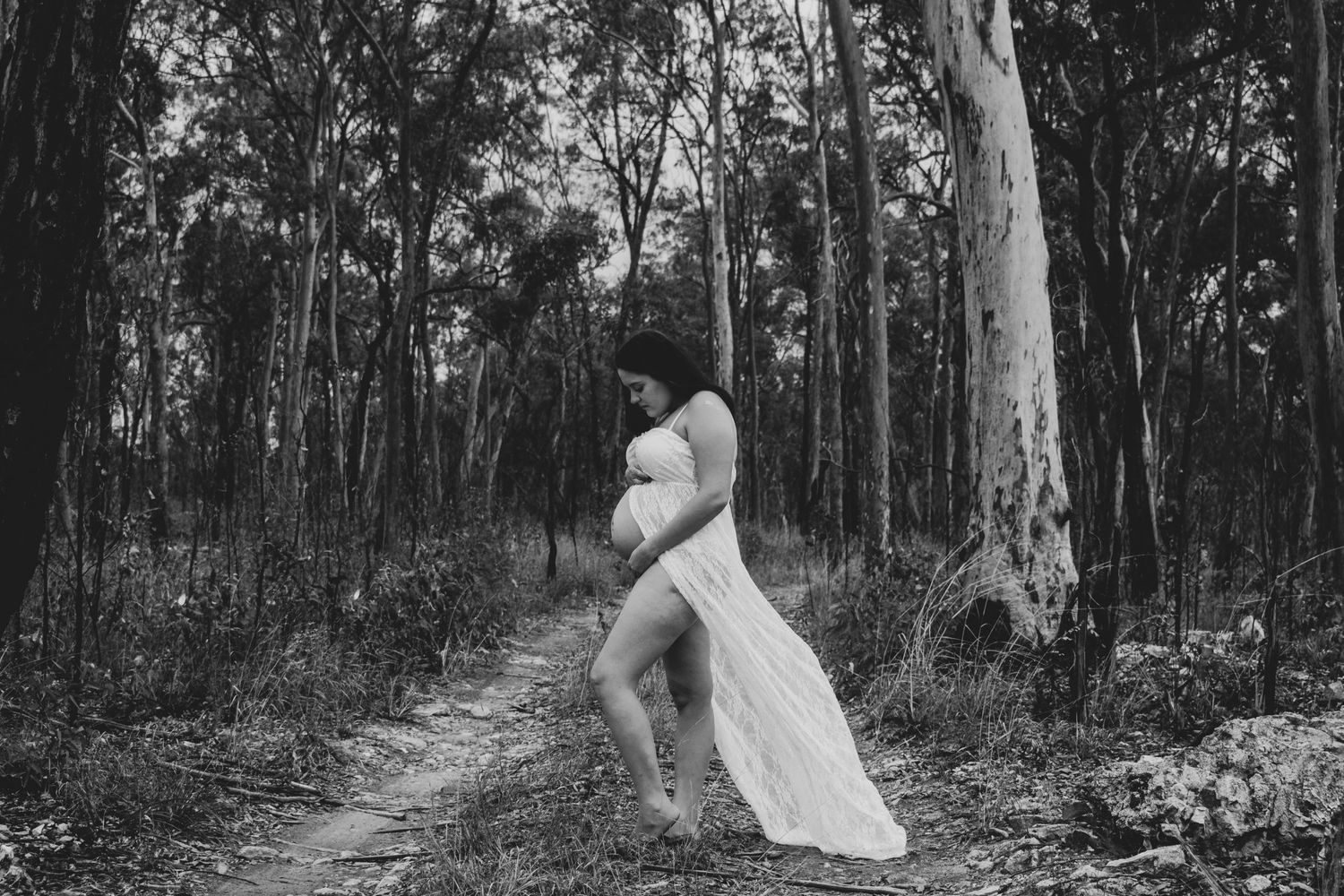 Silhouette of an expectant mother in a flowing dress standing in a forest path.