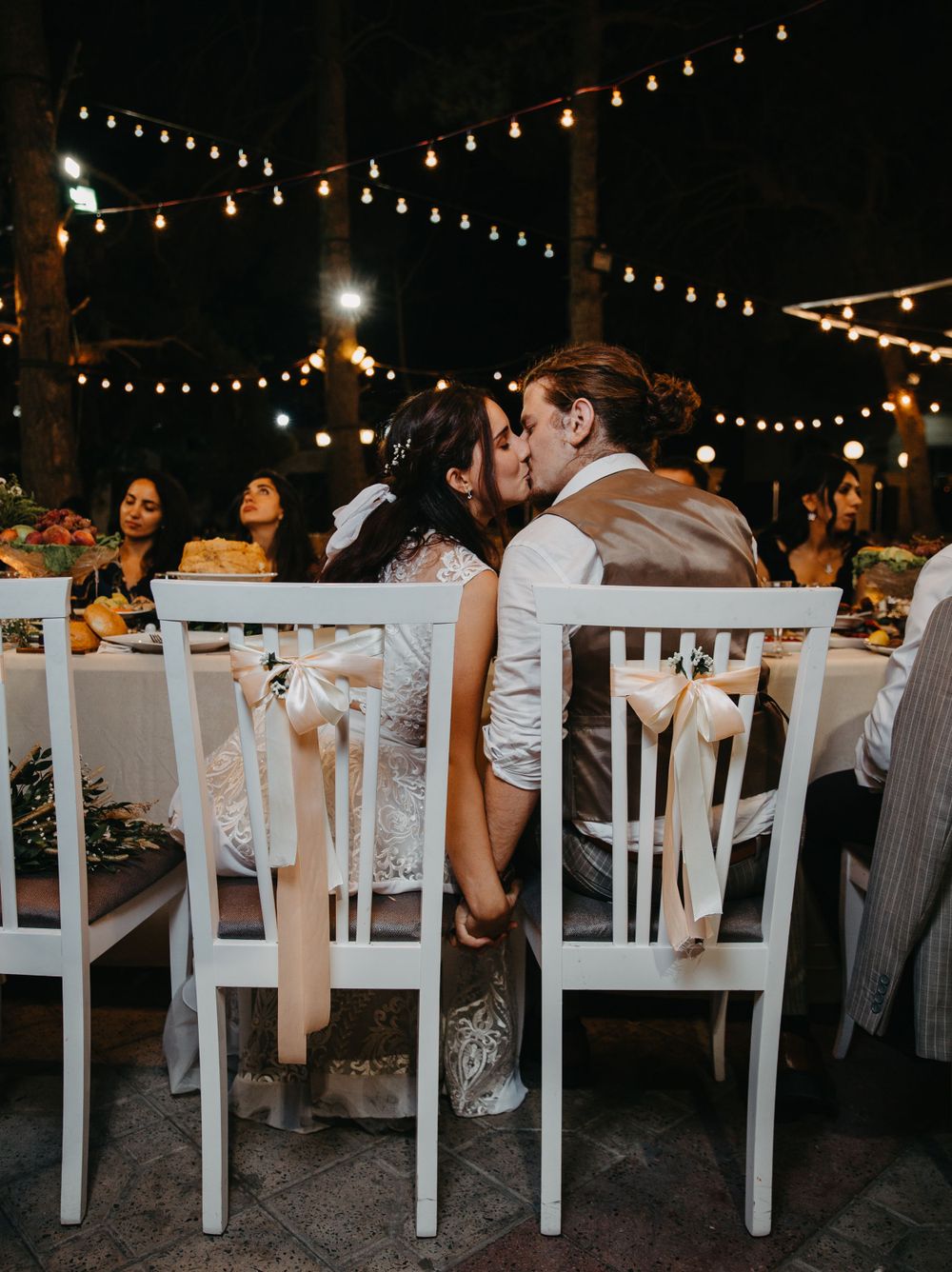 Romantic outdoor wedding reception under twinkling string lights with couple sharing a kiss.