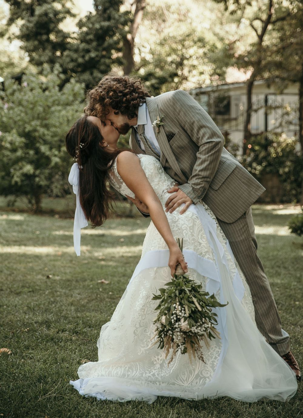 Newlyweds sharing romantic kiss outdoors while bride holds elegant greenery bouquet.