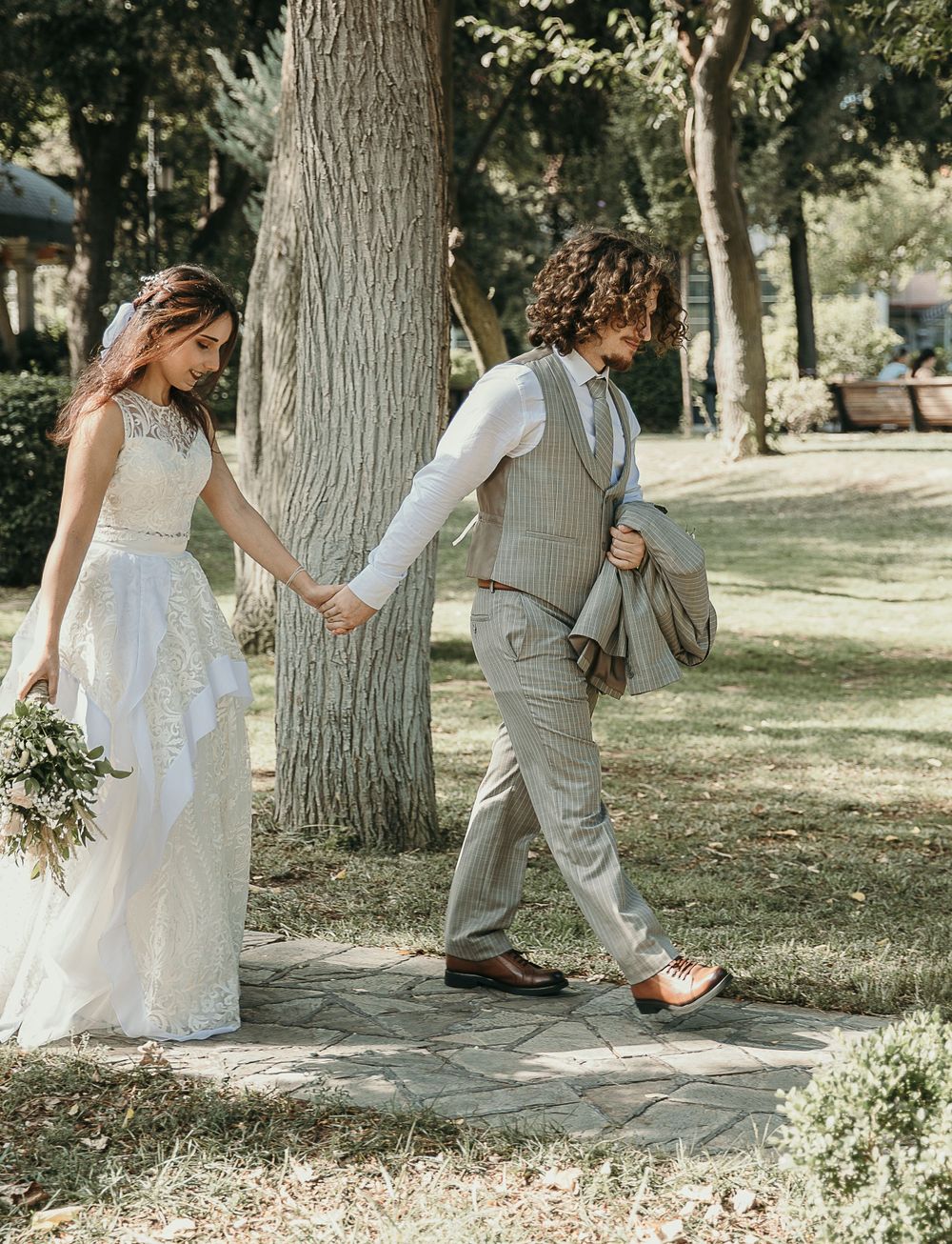 Bride in lace gown and groom in gray vest walking hand-in-hand through park.