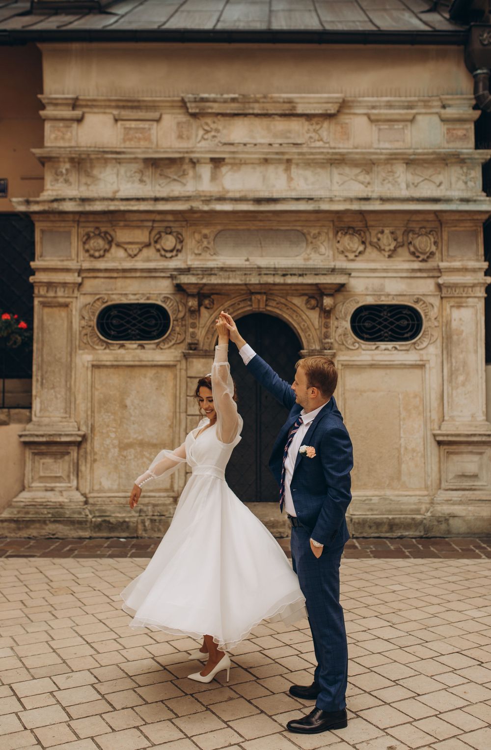 A dancing couple in wedding attire twirls on a stone plaza in front of historic architecture.
