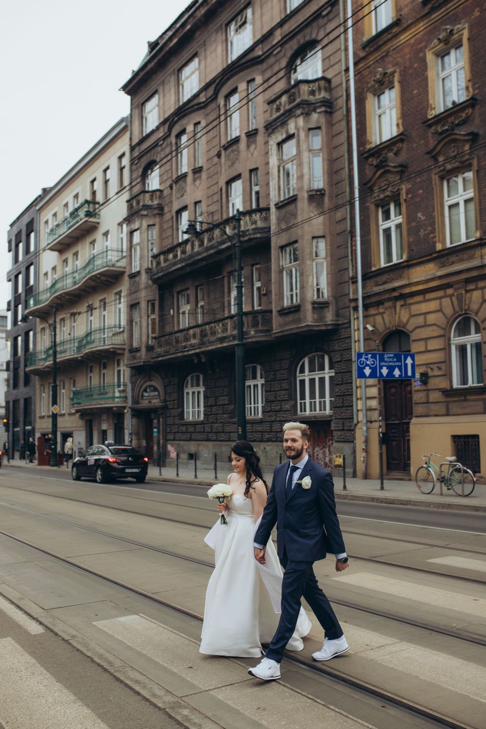 Couple in wedding attire walking on tram tracks against backdrop of historic European buildings.