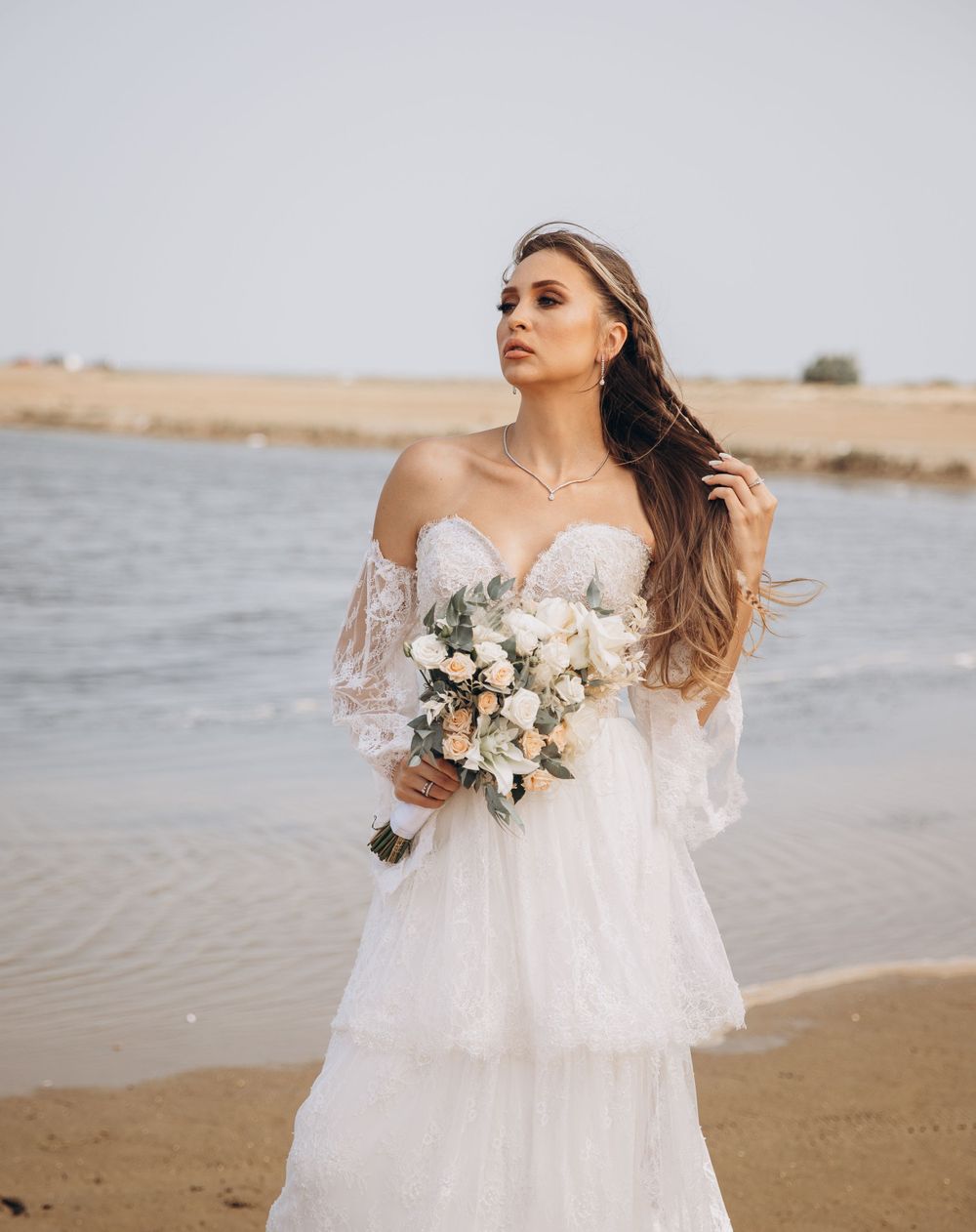A dreamy bridal portrait on the beach featuring a flowing lace dress and neutral bouquet.