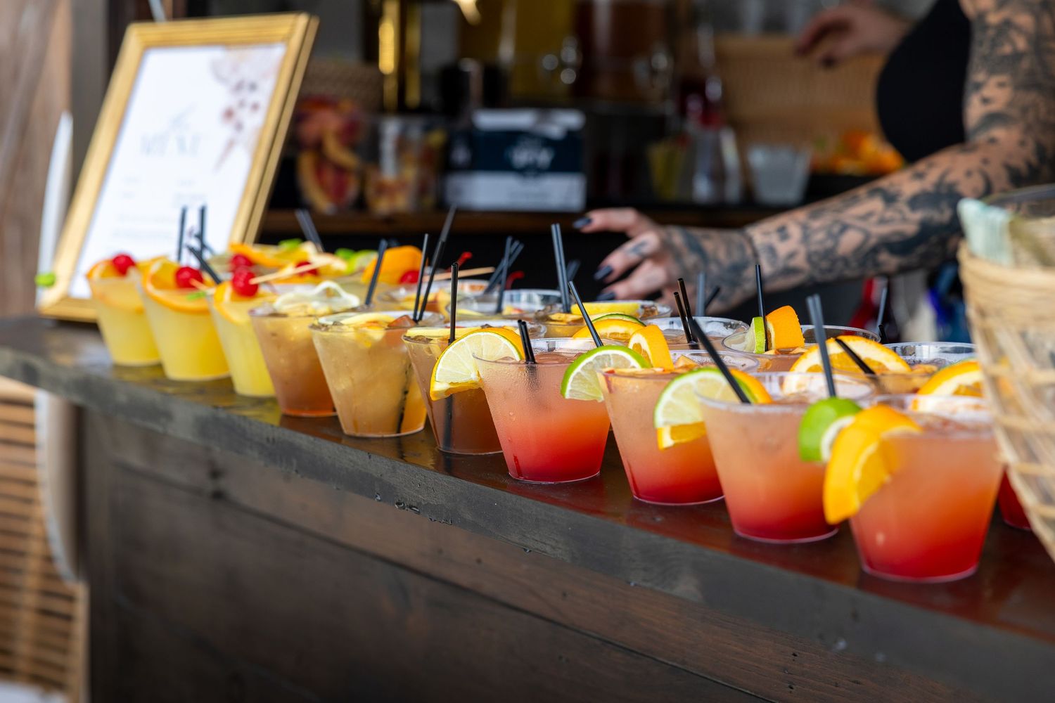 Colorful wedding cocktails lined up on bar with orange and yellow drinks in disposable cups.