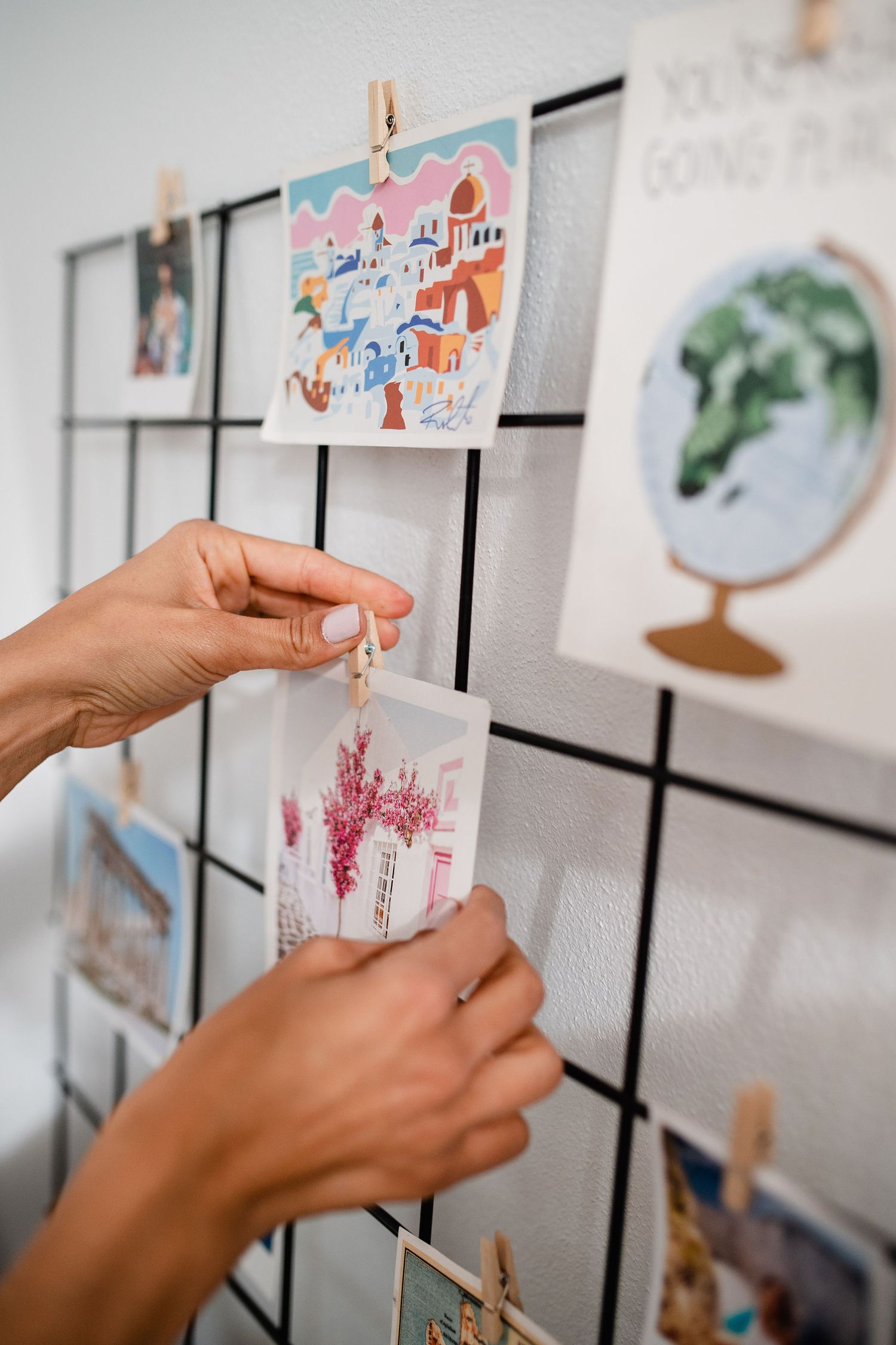 Hands pin colorful artwork featuring houses and landscapes to a wire display grid.