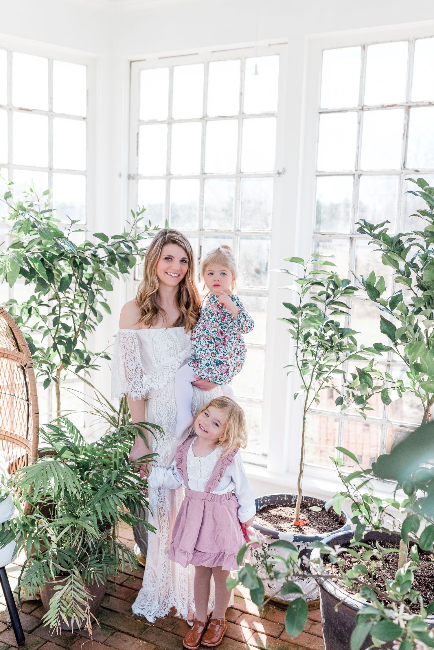A mom and daughters in bright sunlit room with potted plants and windows creating a warm, inviting greenhouse atmosphere.