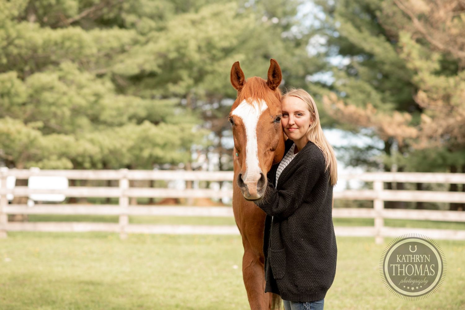 A chestnut horse with white blaze poses with companion in front of wooden fence and pine trees.