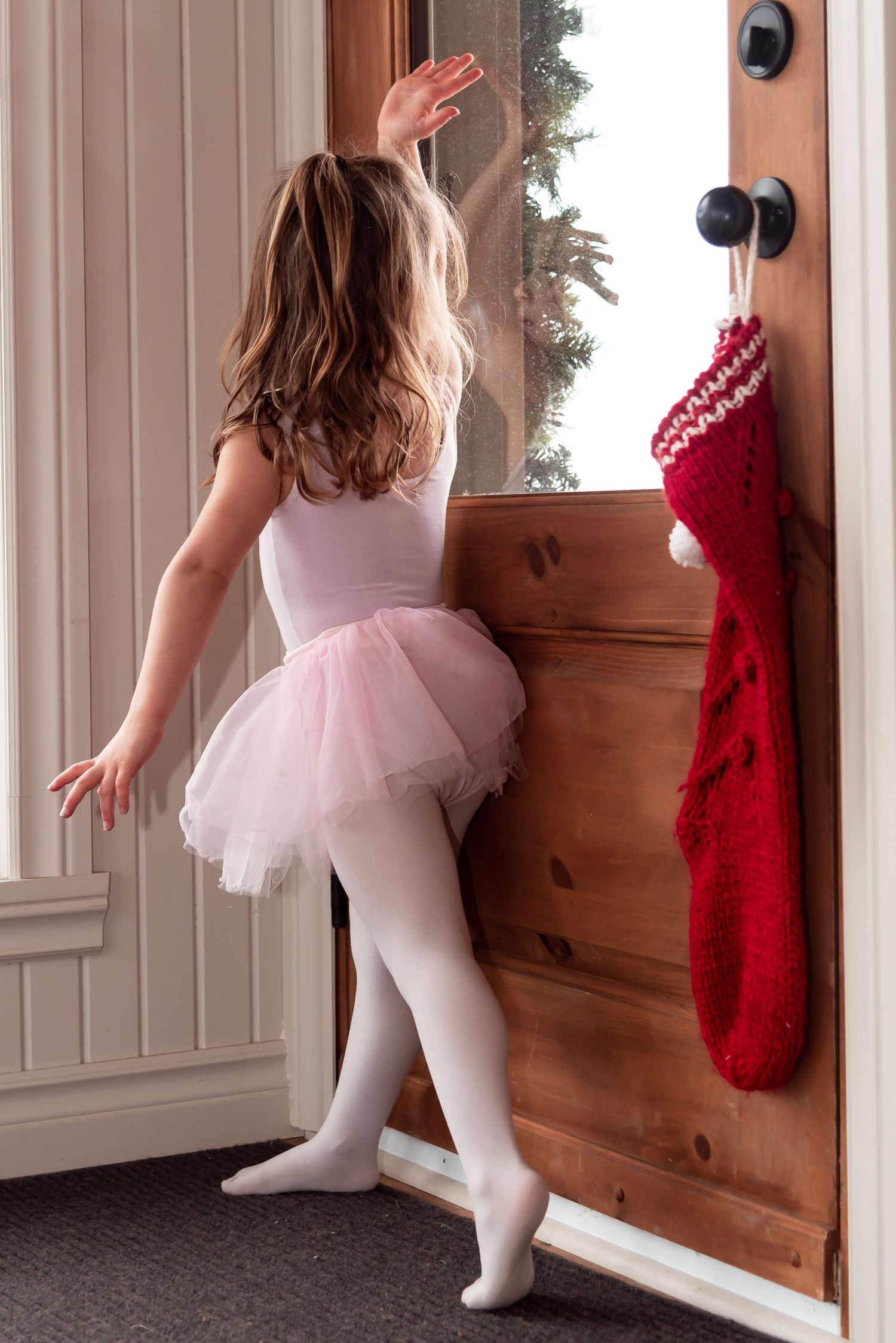 Young ballet dancer in pink tutu and white tights stretches near a door with a red Christmas stocking hanging.