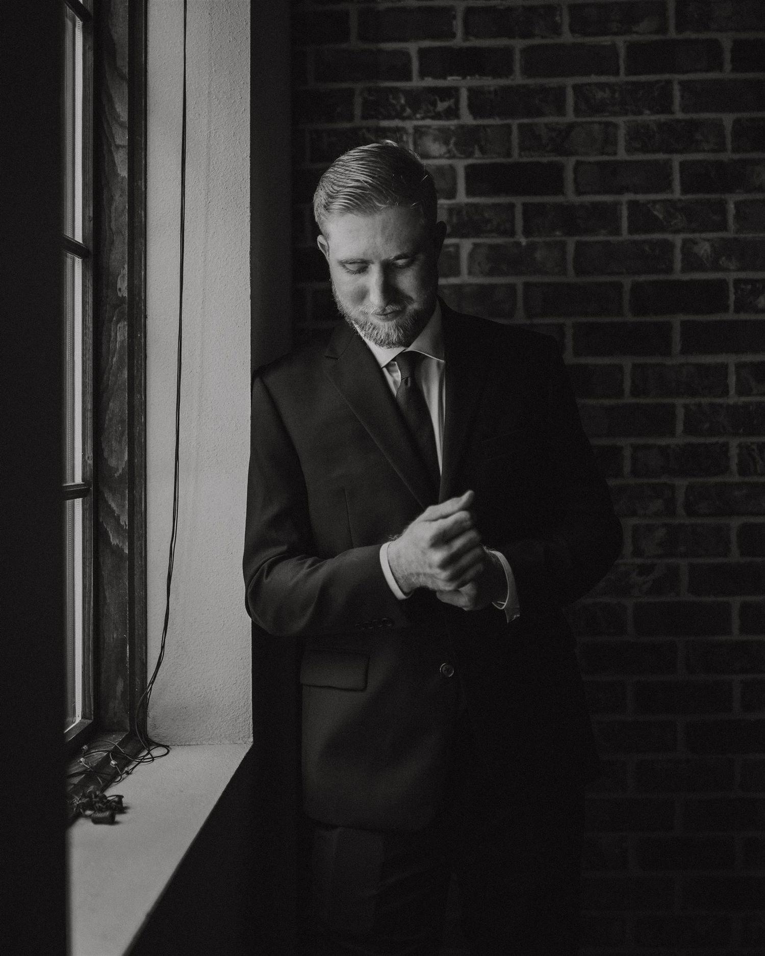 Contemplative figure in suit stands by window with brick wall background in black and white.