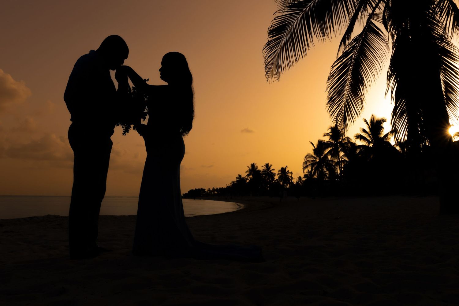 Romantic silhouette of couple at sunset with palm trees and ocean in background.