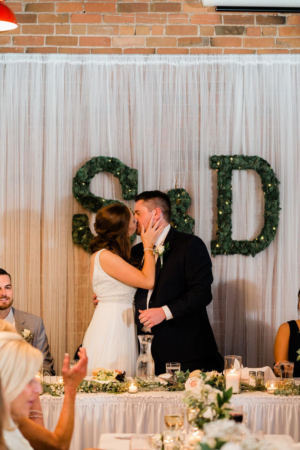Bride and groom kiss at reception head table beneath decorative S and D greenery monogram letters.