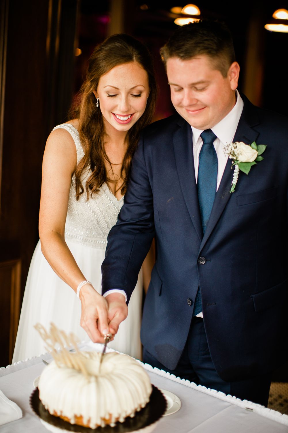 Smiling bride and groom cut their white bundt wedding cake together at the reception.