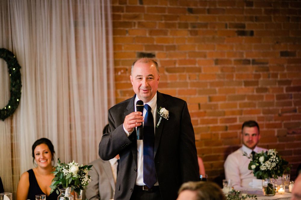 Man in black suit giving a speech at a wedding reception with brick wall backdrop.