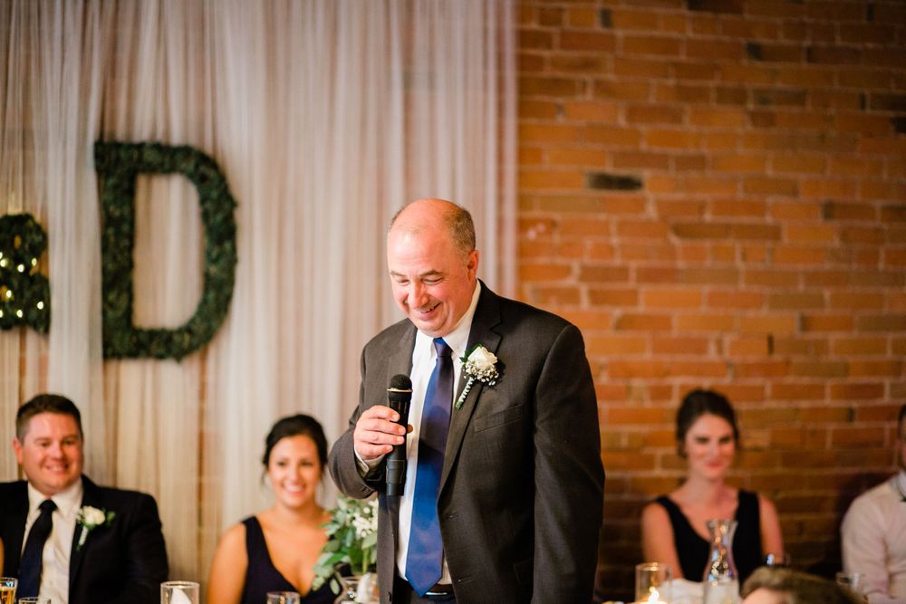 Smiling man delivering wedding speech with greenery letter D decoration behind him.