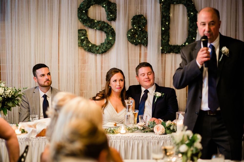 Wedding party at head table with S&D greenery monogram backdrop during reception speech.