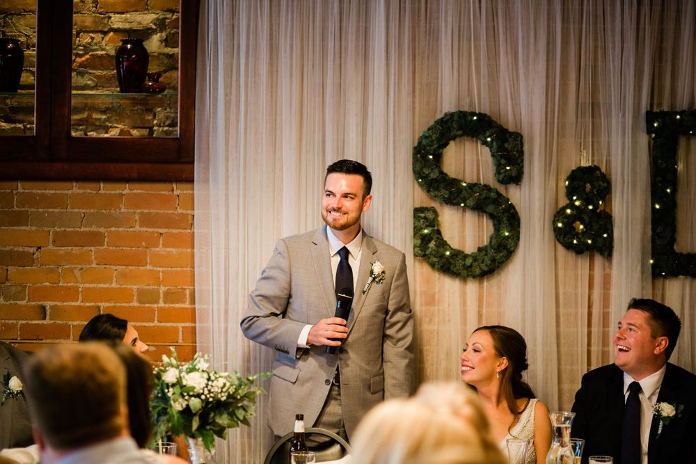 Best man smiling while giving wedding toast with bride laughing in background.