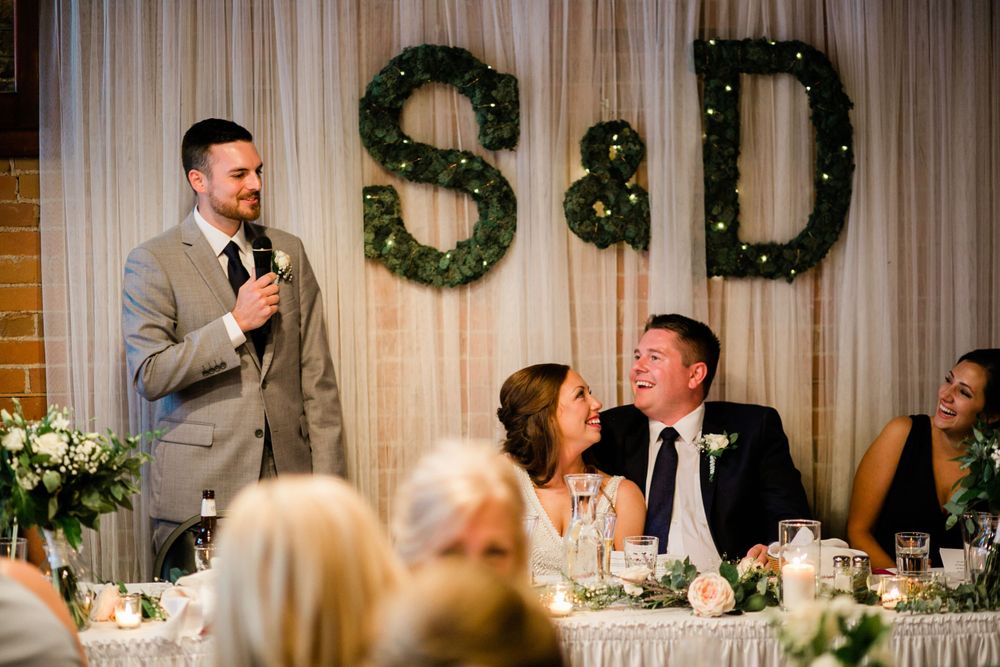 Groomsman giving toast as bride and groom laugh at wedding reception with S&D backdrop.