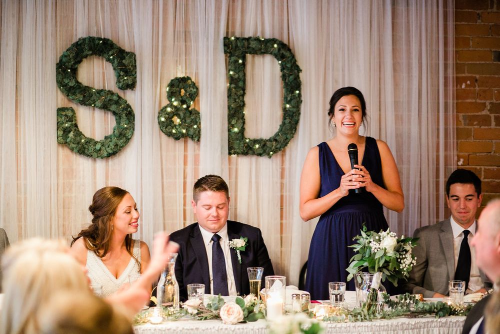 Bridesmaid delivering wedding toast in navy dress with S&D greenery letters behind her.