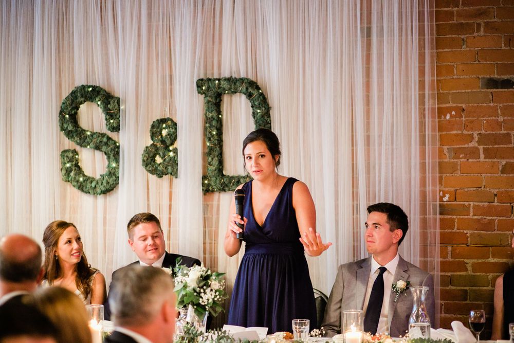Woman in navy dress giving heartfelt speech at wedding reception with S&D monogram backdrop.