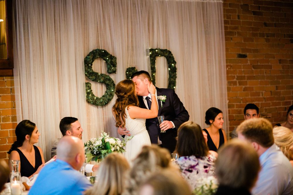 Bride and groom sharing romantic kiss at wedding reception table with S&D monogram backdrop.
