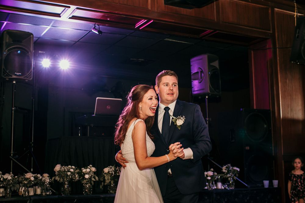 Smiling bride and groom share first dance at elegant wedding reception with purple lighting.
