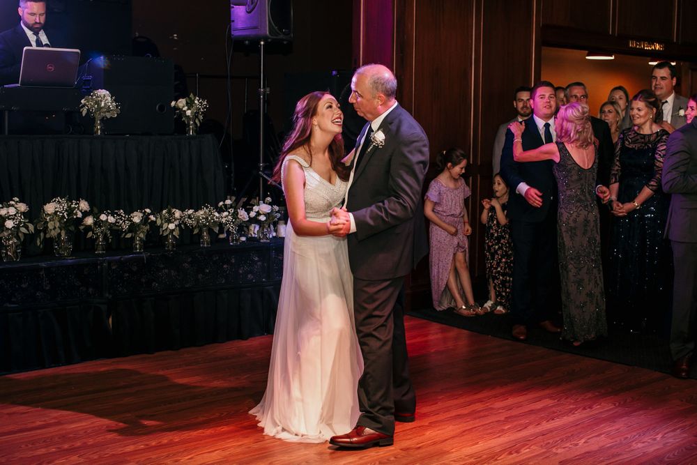Bride shares father-daughter dance at wedding reception as guests watch in the background.