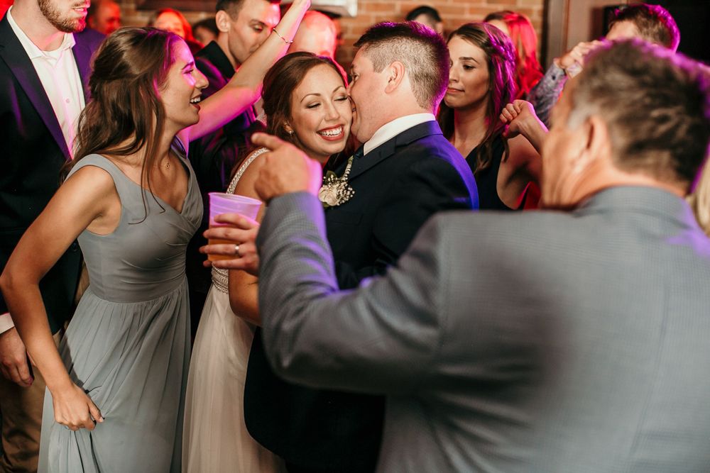 Bride in white gown dances joyfully with wedding guests on colorful reception dance floor.