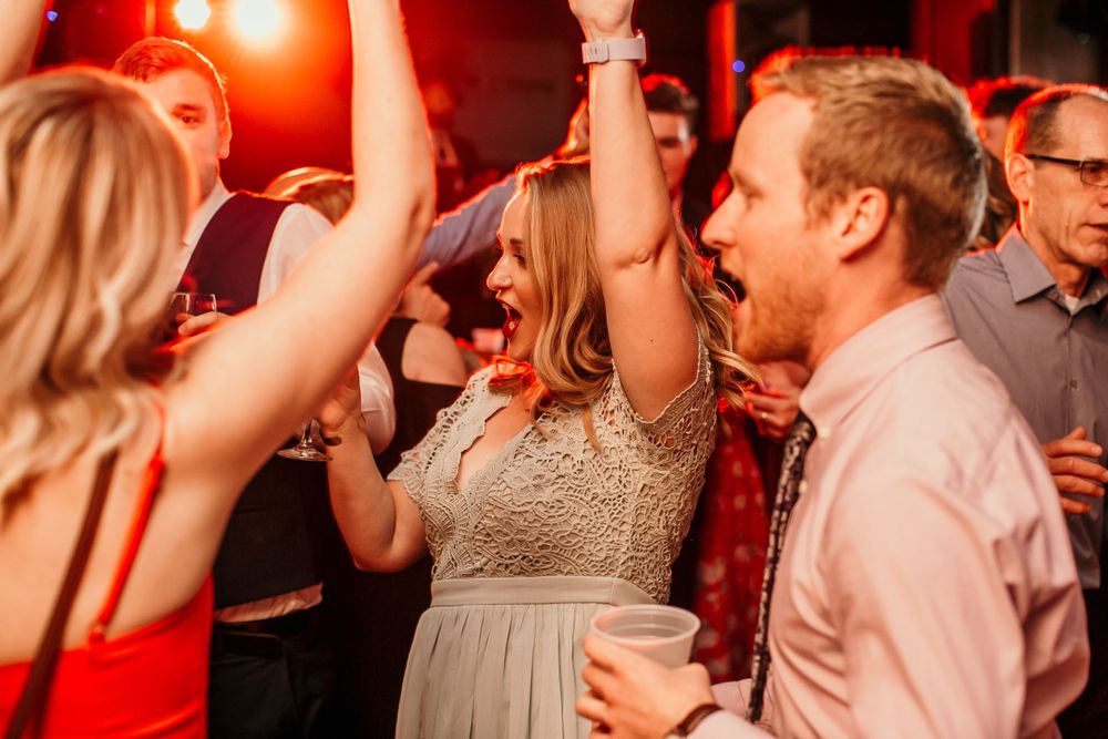 Excited wedding guests dance energetically with raised arms under red lights at reception.