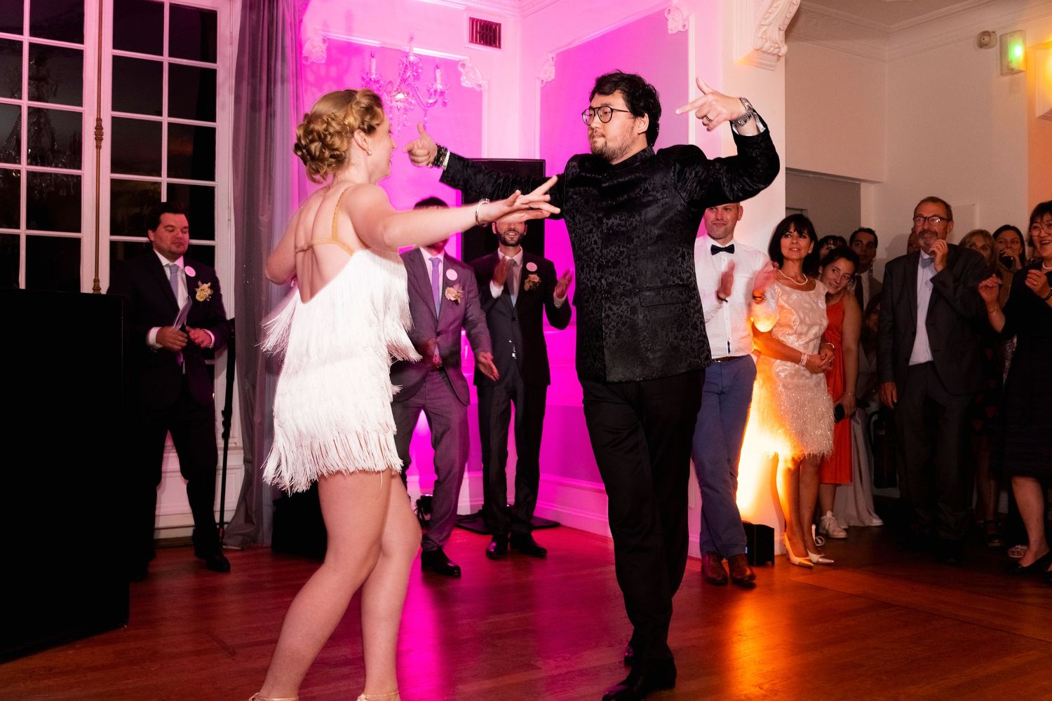 Dancing couple performs energetically in front of wedding guests under pink lighting.