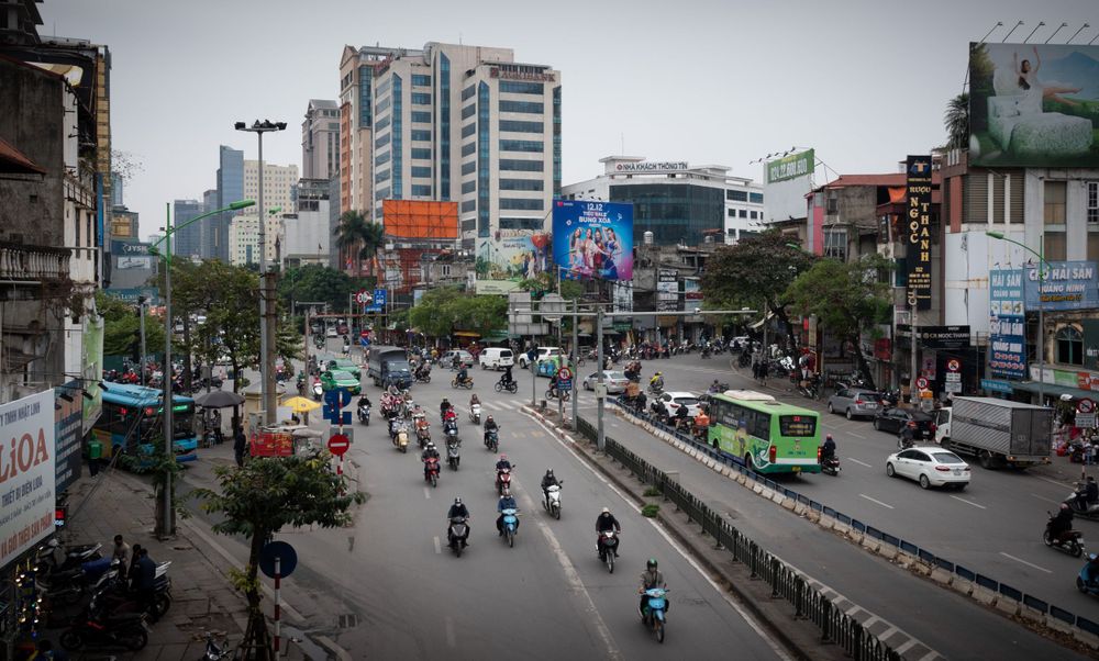 Wide urban street view with motorcycles and vehicles traveling past modern city buildings.