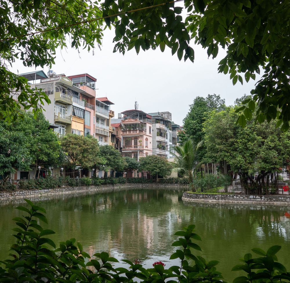 Residential buildings reflect in a calm lake surrounded by leafy green trees.