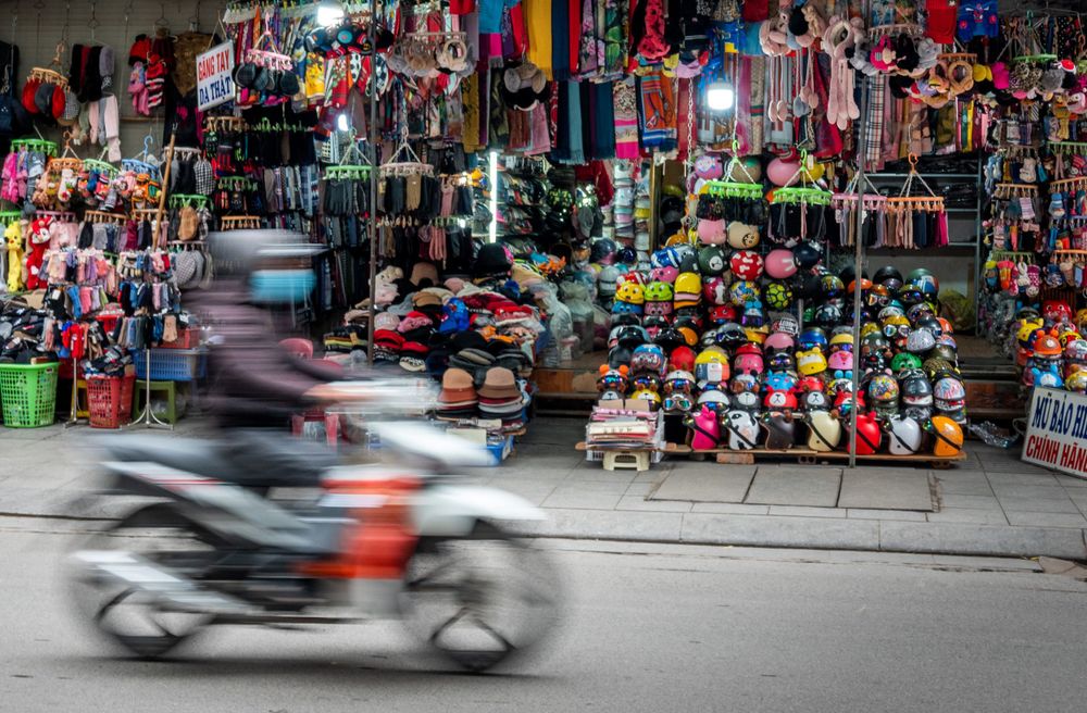 Motion-blurred motorcycle passing by a vibrant storefront display of helmets and clothing in Vietnam.