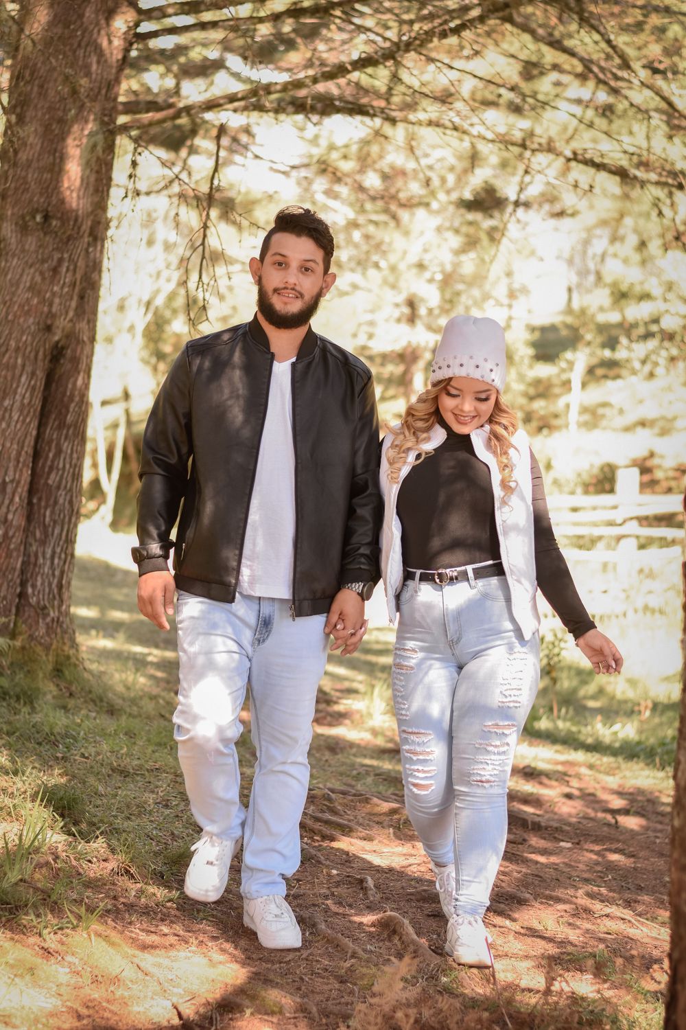 A couple in matching leather jackets and ripped jeans walks together on a sunlit forest path.