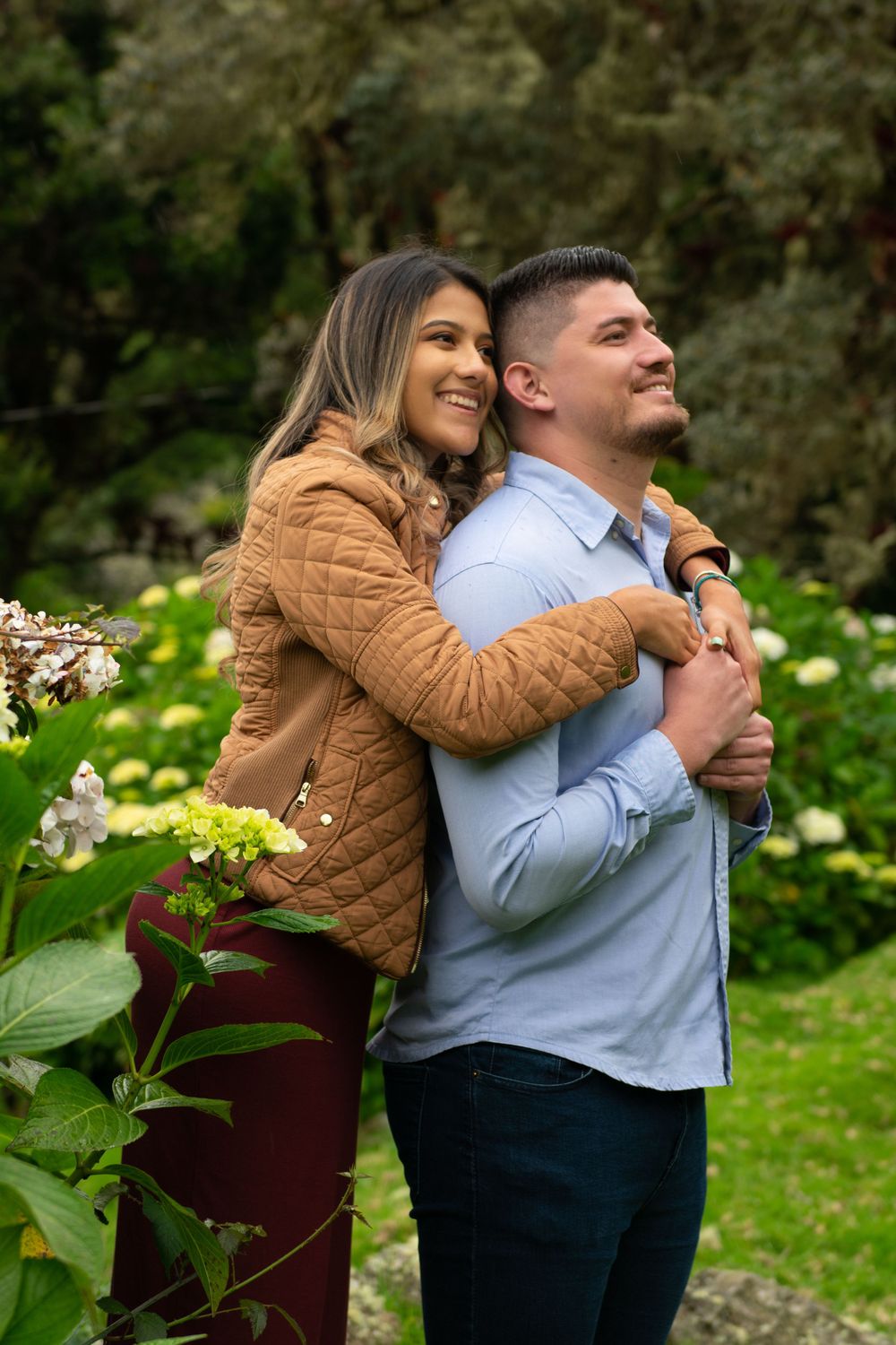 Couple embracing in garden setting surrounded by blooming flowers and greenery.