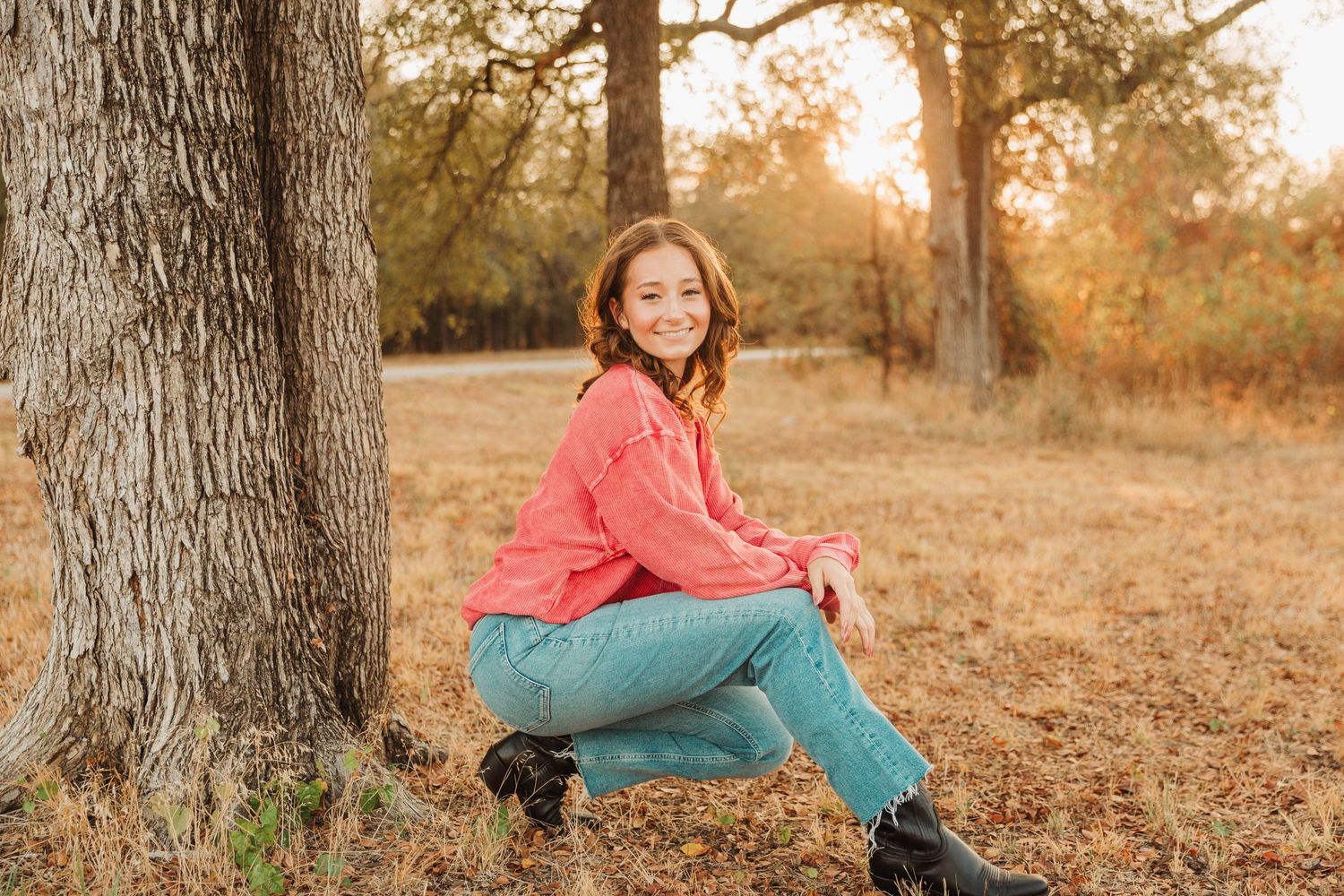 Senior girl in coral sweater sits by tree trunk in autumn field at sunset.
