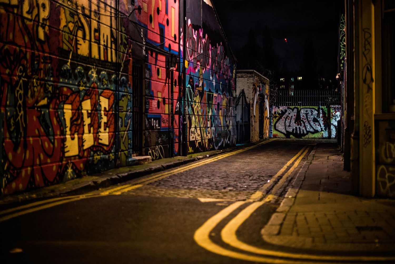 Colorful graffiti adorns walls along a dark urban alleyway with yellow street lines.