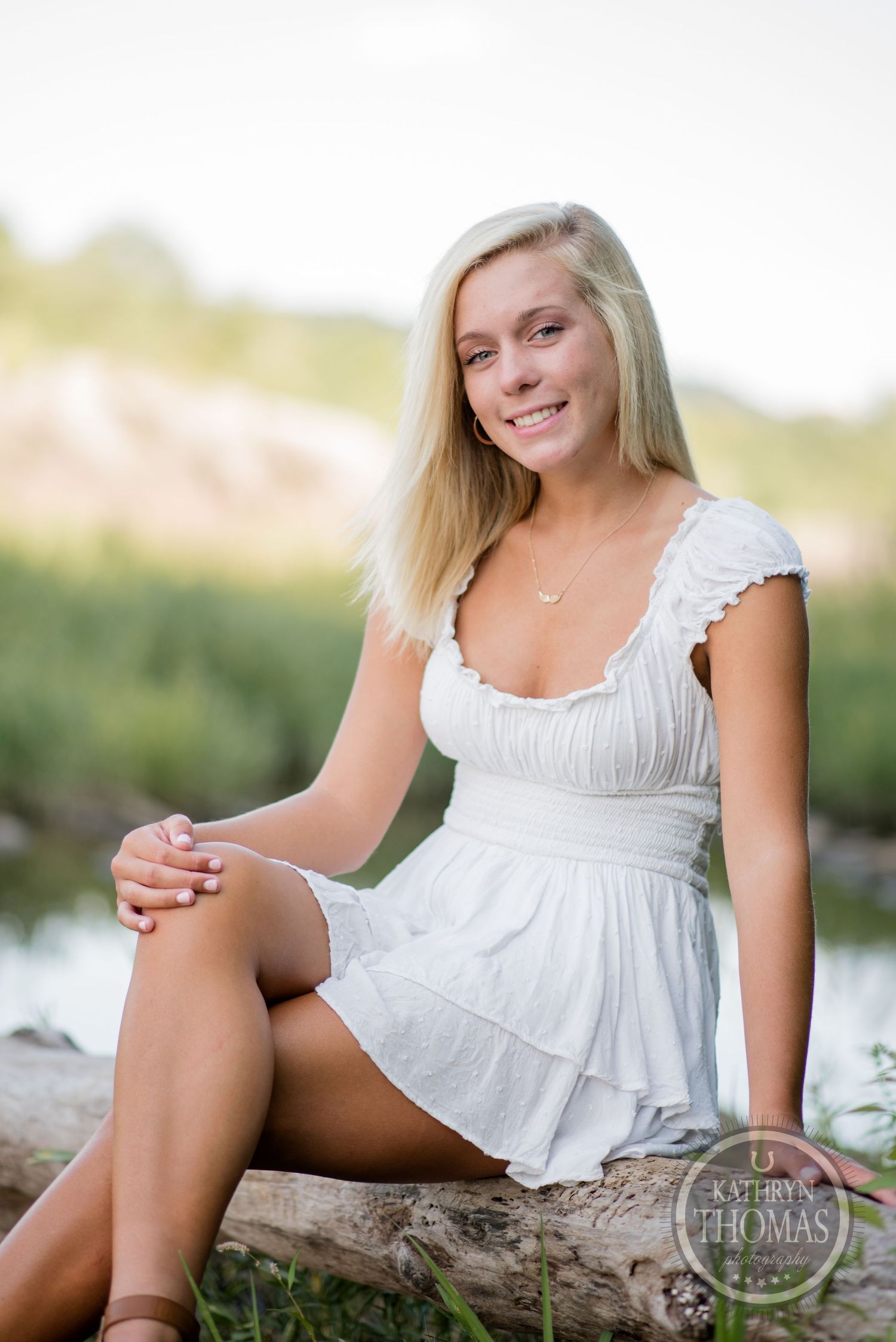 Woman in white sundress sitting casually on log by water.