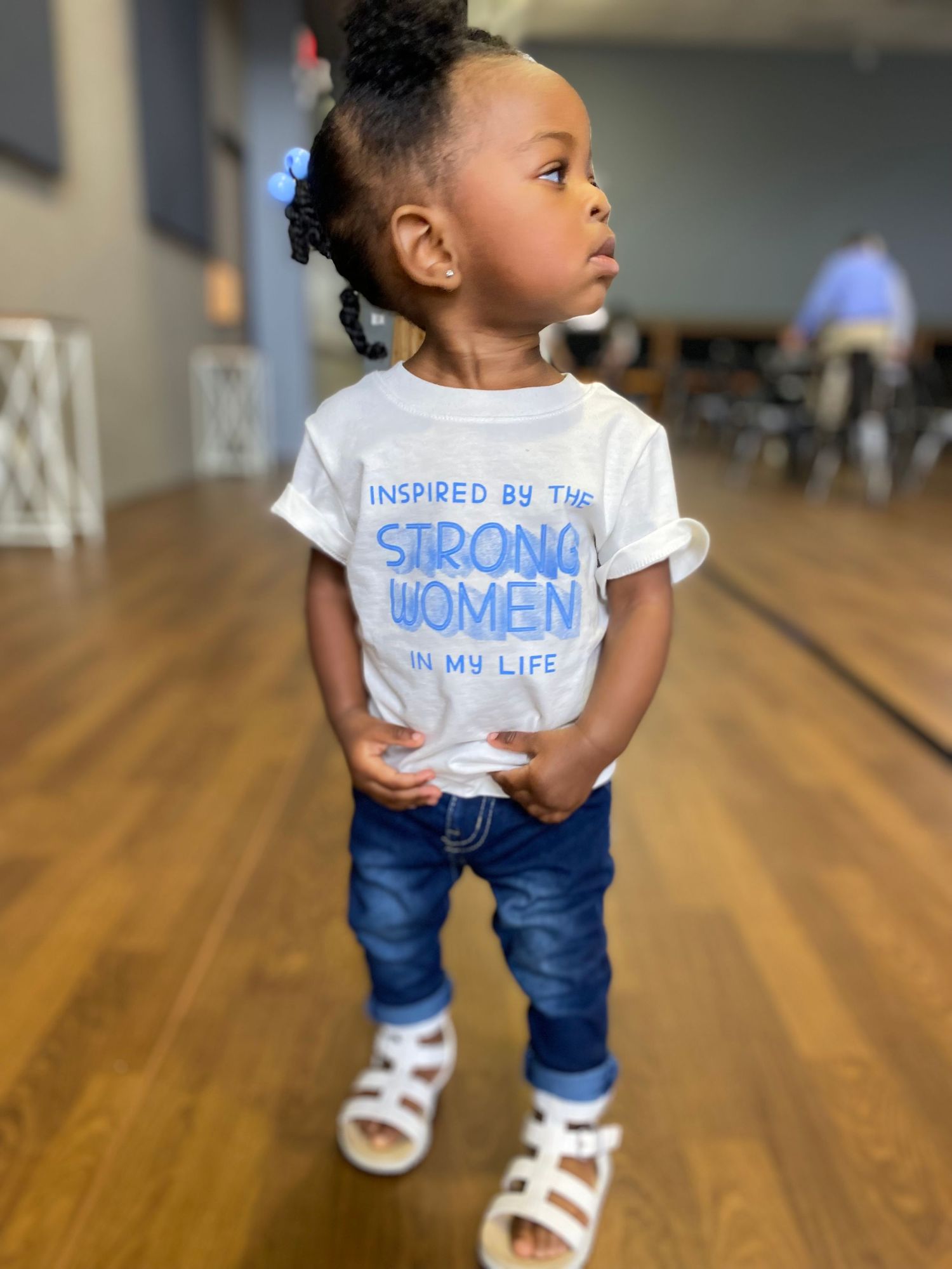 Young child wearing t-shirt with empowering message about strong women on hardwood floor.