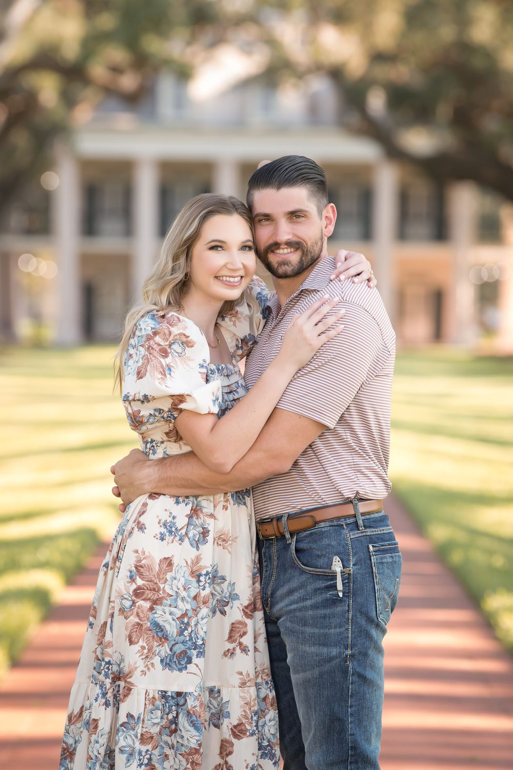A romantic photoshoot on a tree-lined pathway with a white colonial building visible in the distance.