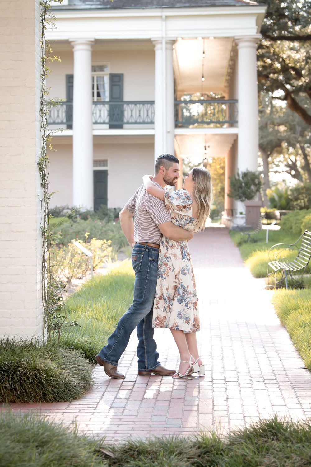 Engagement photos taken on a brick walkway with white pillars and landscaping in the background.
