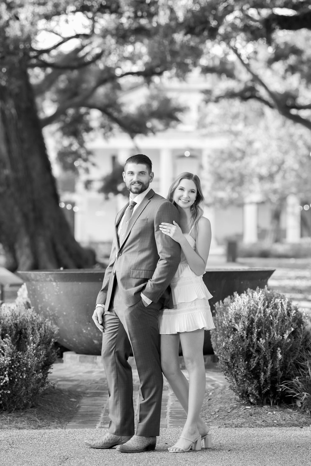 Black and white engagement photos taken near decorative shrubs and trees.